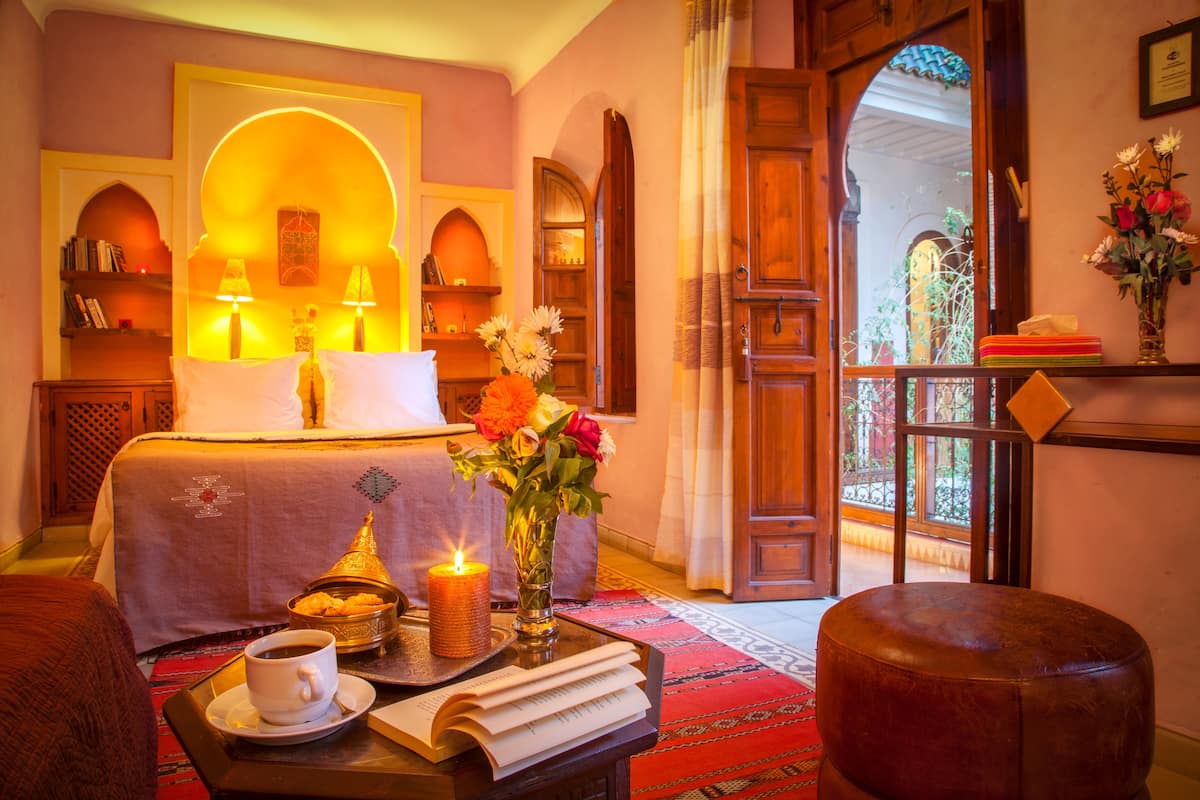 A room with a bed, a table with a cup of coffee, a candle, and a book.