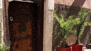 A rustic wooden door with a metal plaque, a small wall-mounted light fixture, and potted plants on a cobblestone path. - Riad Dar Nabila (Marrakech)