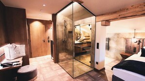 A modern bedroom with a large bed, a wooden bench, a glass-enclosed shower, and a wooden ceiling beam. - Antonius (Kaprun)