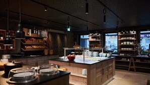 A modern kitchen with wooden cabinetry, a central island with a buffet setup, and a view of the street through large windows. - Antonius (Kaprun)