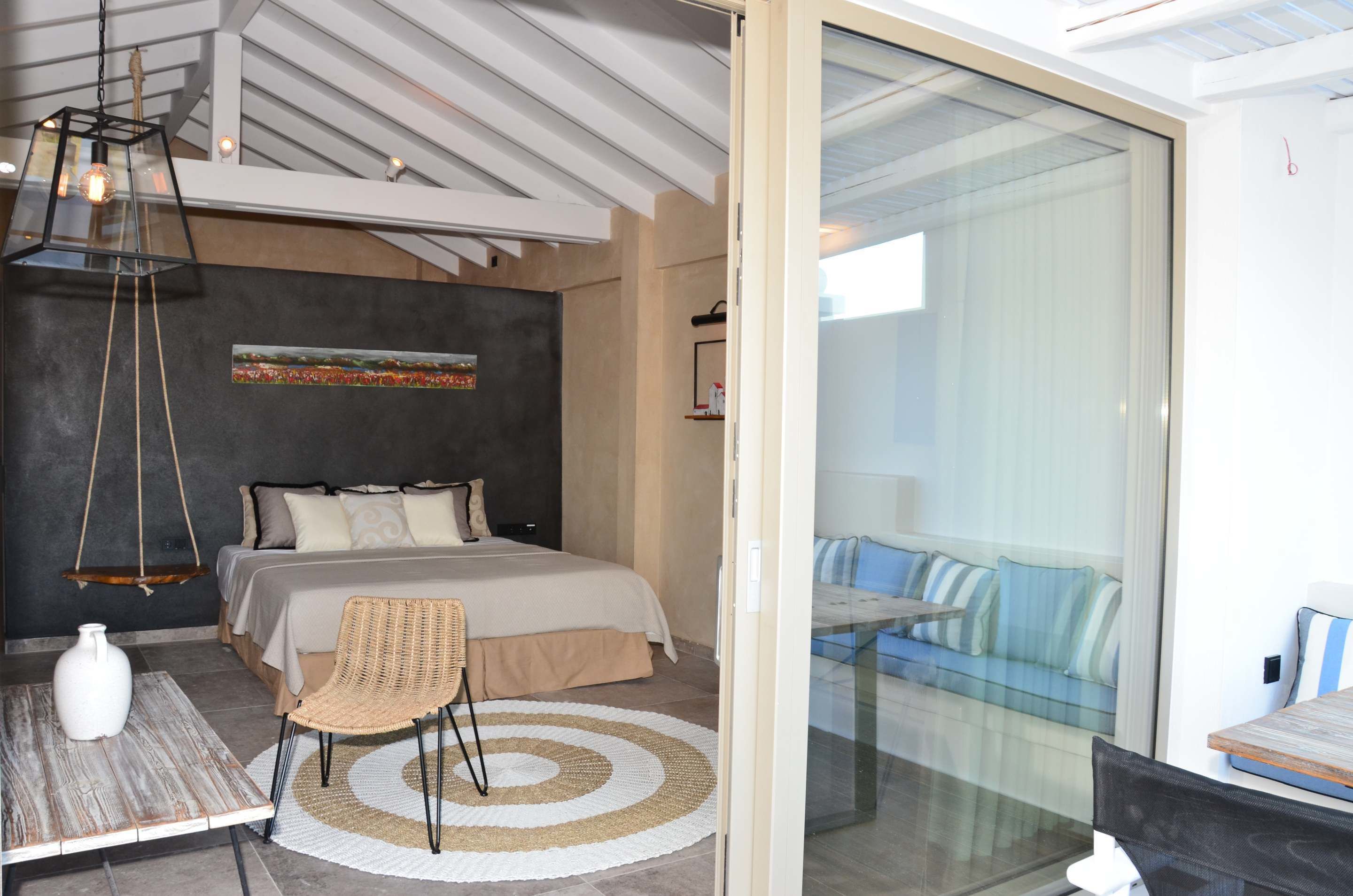 A modern bedroom with a bed, a hanging light fixture, a wicker chair, and a glass door leading to a balcony with a sofa.