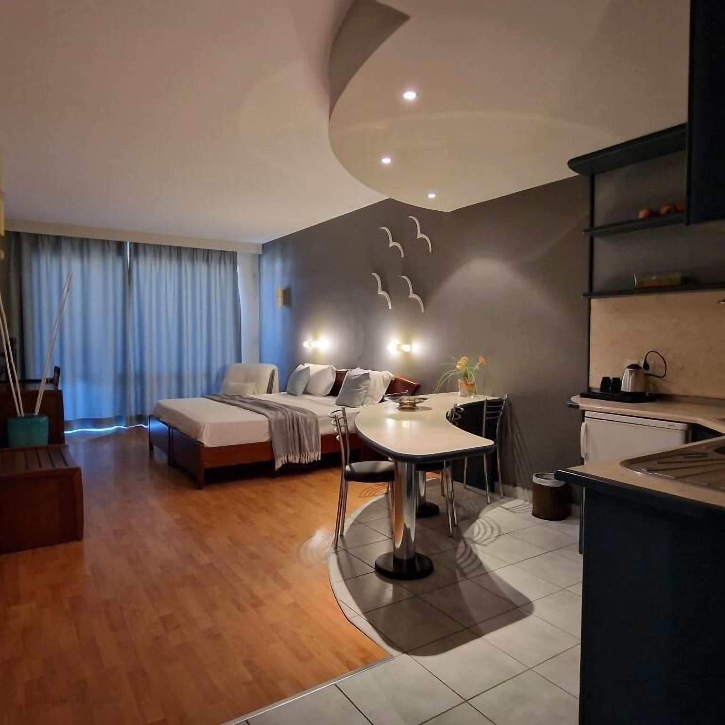 A modern hotel room with a bed, a dining area, and a kitchenette.