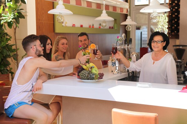 A group of people toasting with drinks at a bar with a fruit platter. - Evilion Sea And Sun (Dio-Olympos)