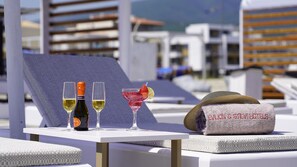 A table with two glasses of white wine, a bottle of wine, a pink cocktail with a strawberry and lime, and a towel with "EVION & STELV hotel's" embroidered on it. - Evilion Sea And Sun (Dio-Olympos)