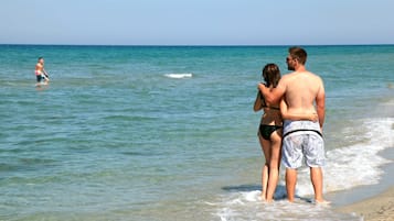 Une scène de plage où l’on voit un couple debout sur le rivage, une personne nageant dans l’eau et des vagues qui viennent doucement se briser sur le sable.