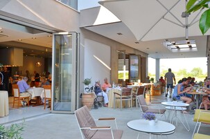 Un área de comedor al aire libre con mesas y sillas, una entrada con puerta de vidrio y vistas al exterior.
