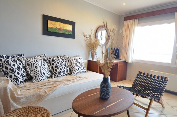 A living room with a sofa, patterned cushions, a round wooden table, a woven chair, a vase with dried plants, and a painting on the wall. - Evilion Sea And Sun (Dio-Olympos)
