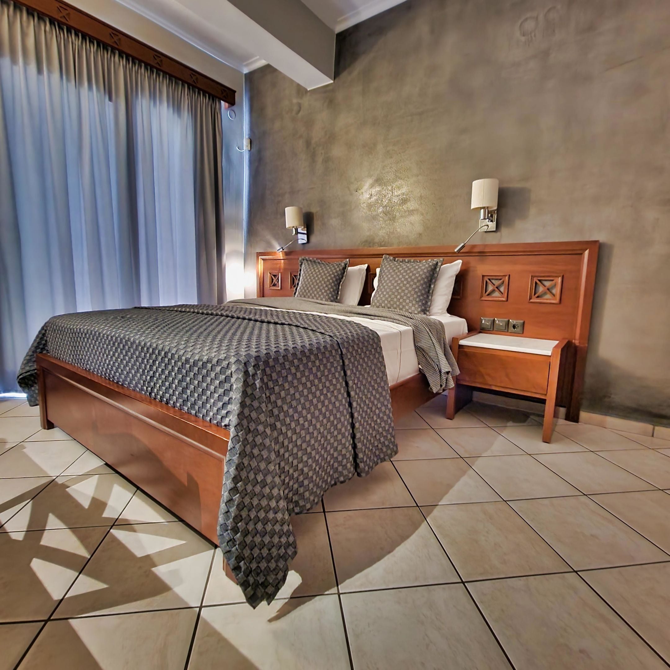 Una habitación de hotel con una cama grande, una mesita de noche, una lámpara de pared y una ventana con cortinas.