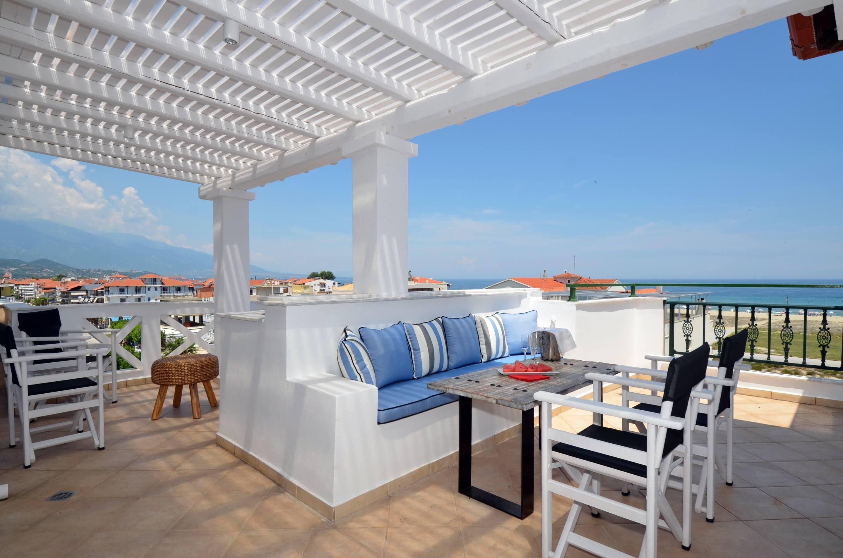 Un espace extérieur aménagé avec des sièges, abrité et offrant une vue sur la mer, avec du mobilier blanc et une table sur laquelle se trouve un objet rouge.