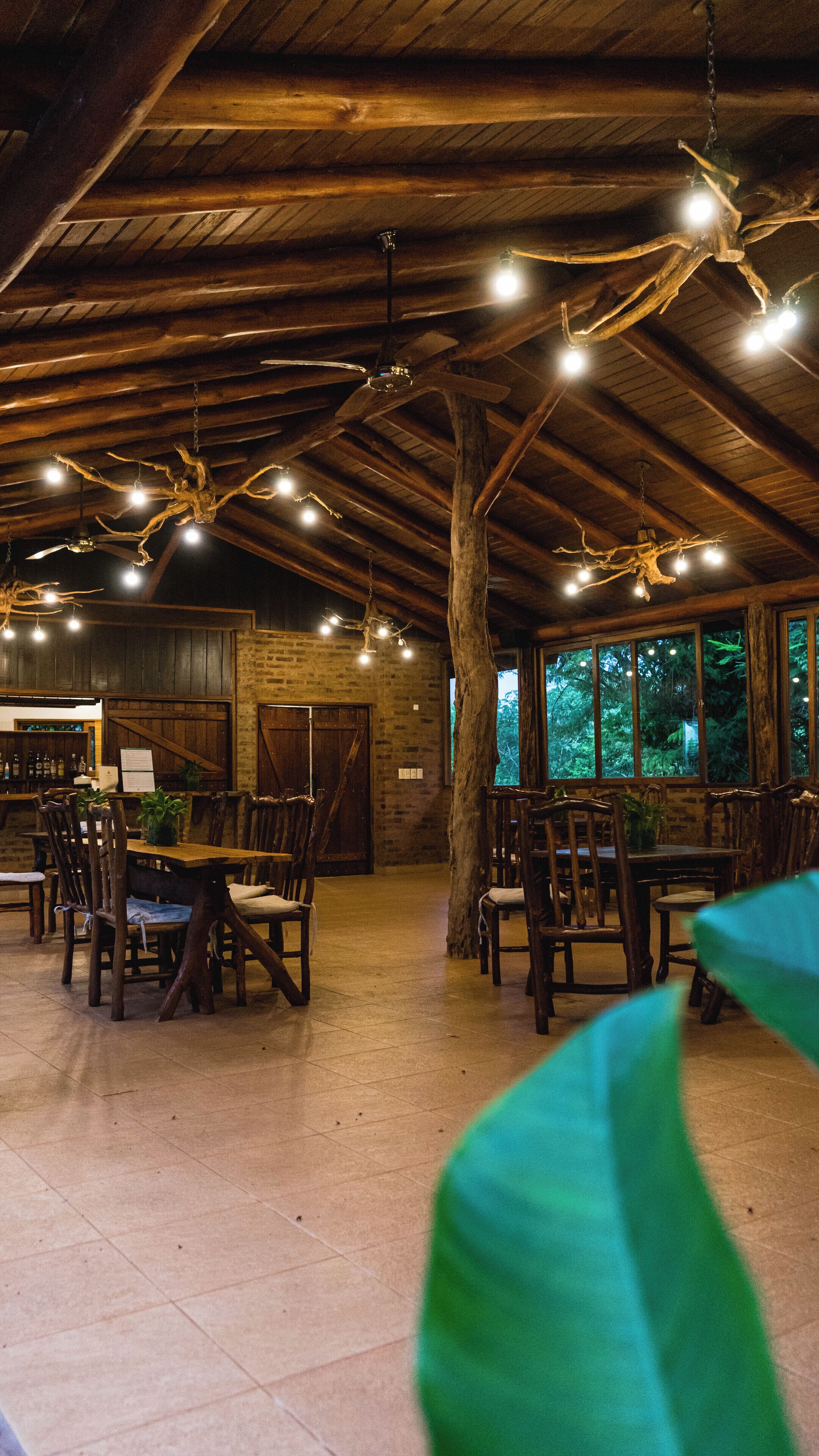 An indoor dining area with wooden tables and chairs, a brick wall, and a large tree trunk in the center.