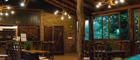 An indoor dining area with wooden tables and chairs, a brick wall, and a large tree trunk in the center.