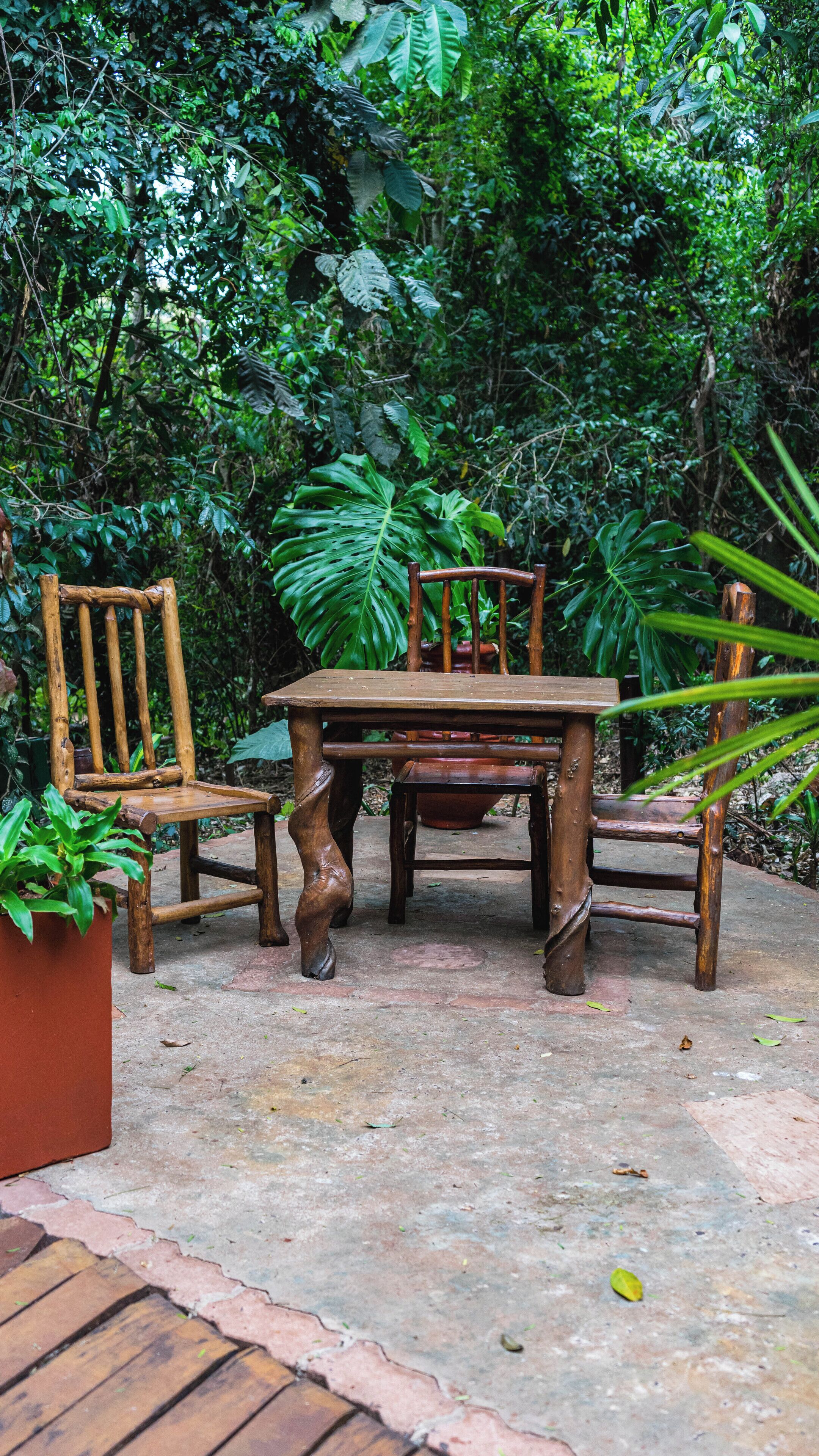 Une table et des chaises en bois sont disposées à l’extérieur, sur un sol en béton, et l’ensemble est entouré d’un feuillage verdoyant et luxuriant.