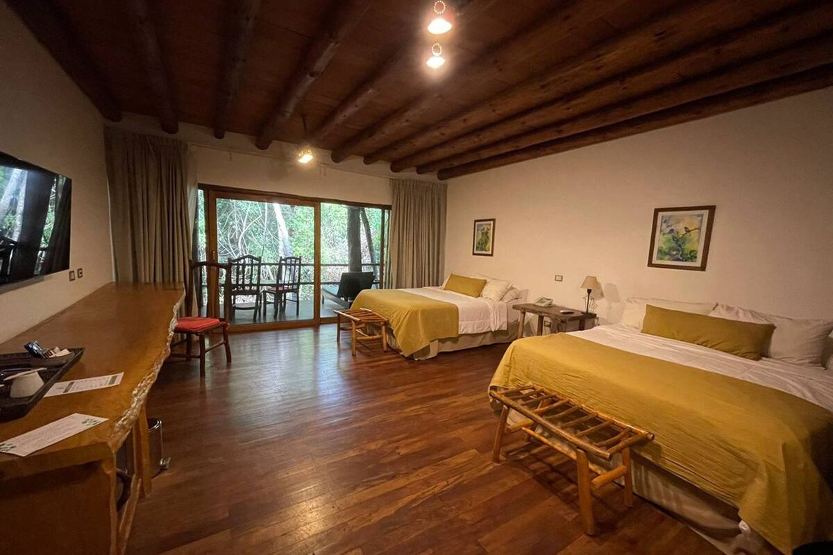 Una habitación de hotel con dos camas, un escritorio de madera, un balcón con sillas y un ventanal grande.