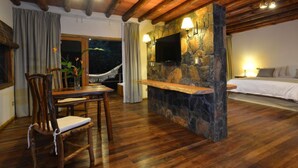 A wooden-floored room with a stone wall, a TV mounted above it, a bed with pillows, and a hammock by the window. - La Aldea de la Selva Lodge (Puerto Iguazú)