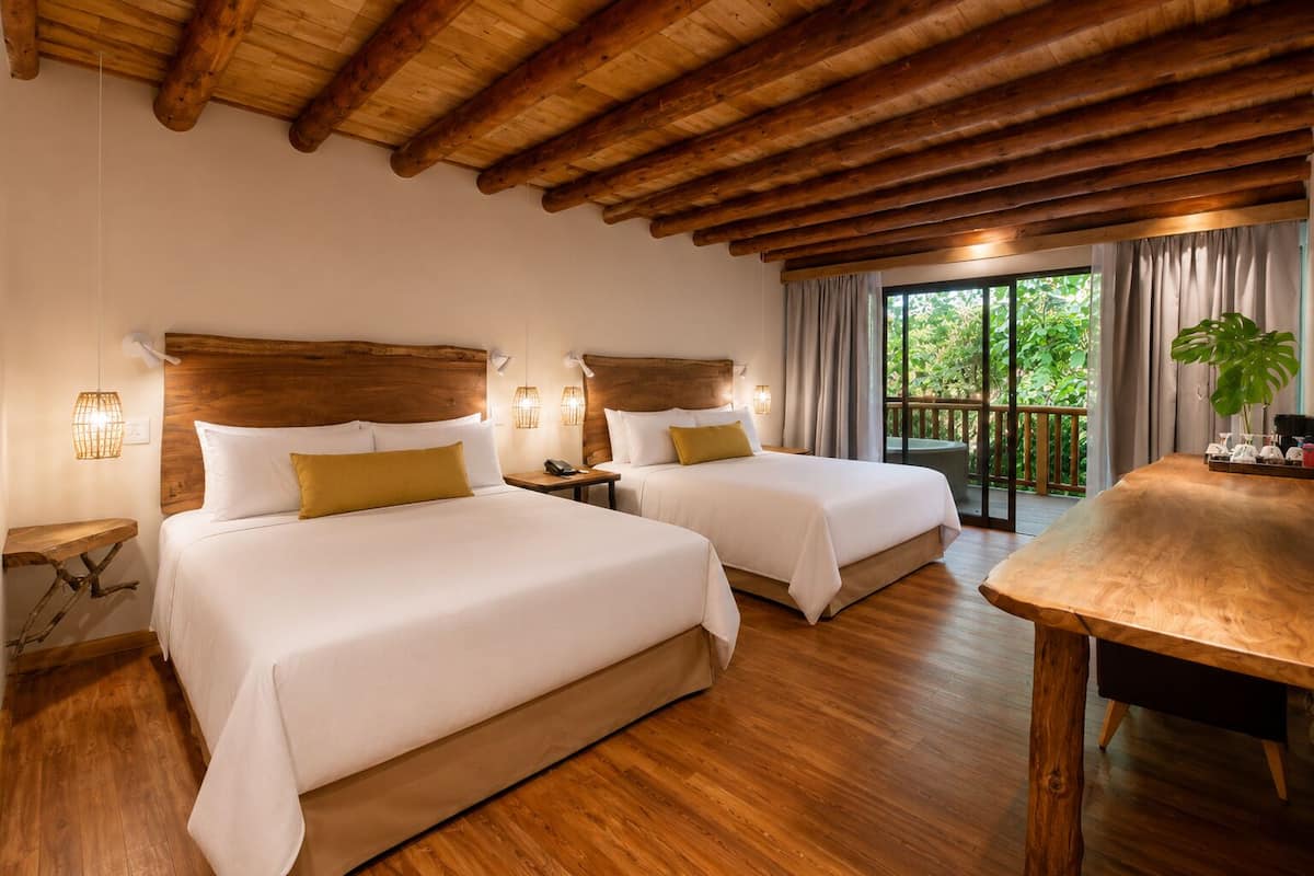 Habitación de hotel con dos camas, suelo de madera, balcón con cortinas y una mesa con una planta.