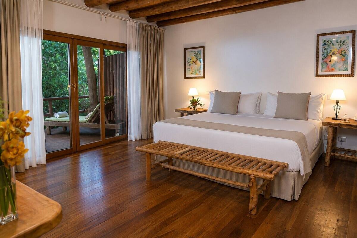 Un dormitorio con una cama grande, suelo de madera, puertas de cristal correderas, dos mesitas de noche con lámparas y cuadros en las paredes.