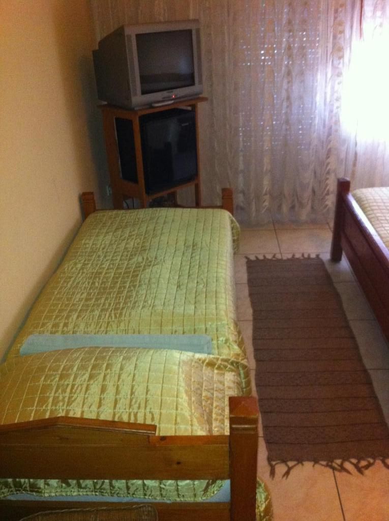 A single bed with a wooden frame and a green bedspread, a television on a stand, and a window with sheer curtains.