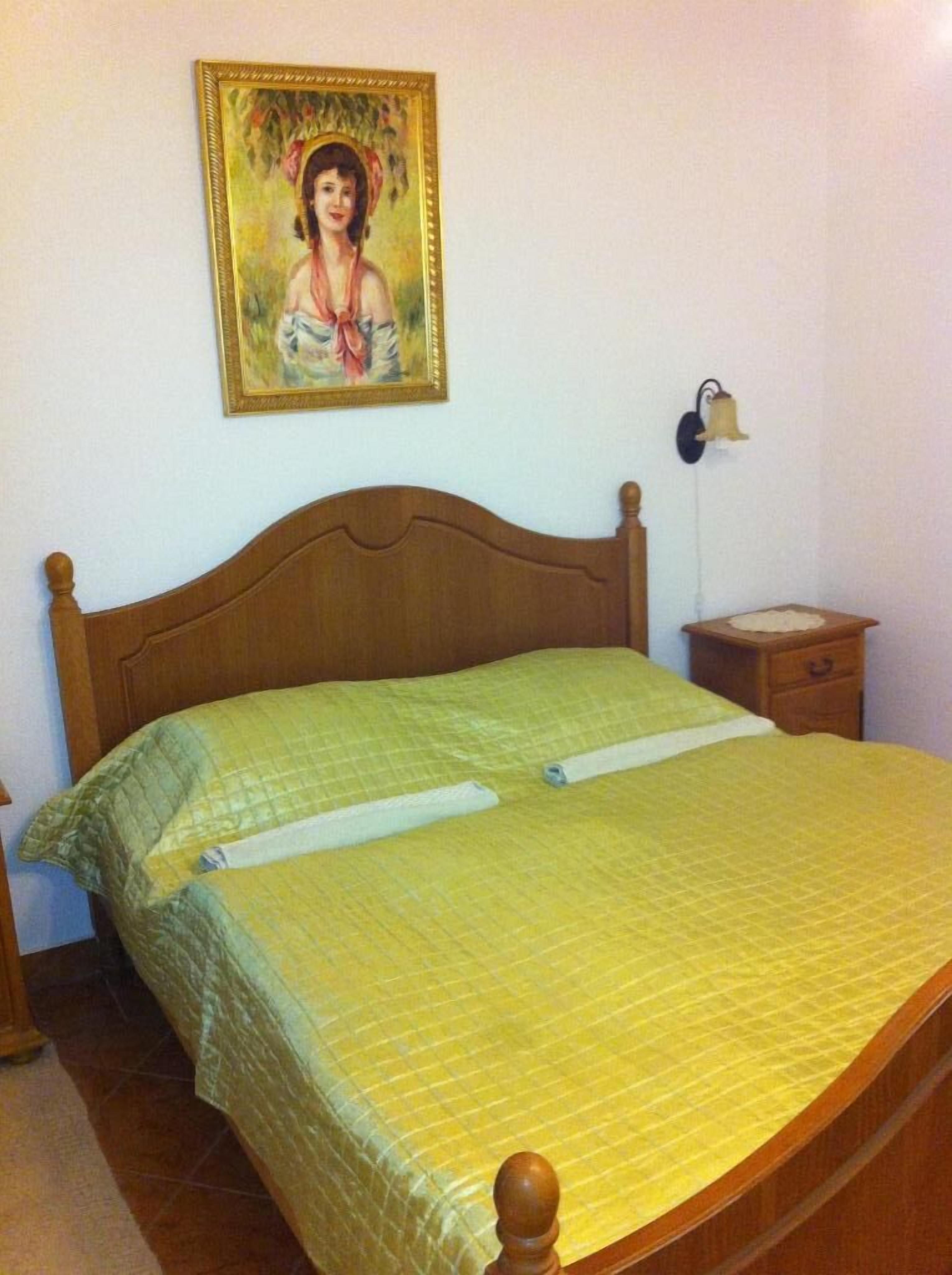 A bedroom with a wooden bed, a nightstand, a wall-mounted lamp, and a framed picture.