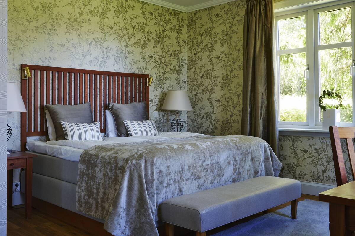 Een slaapkamer met een bed, nachtkastje, lamp, raam en uitzicht op groen.