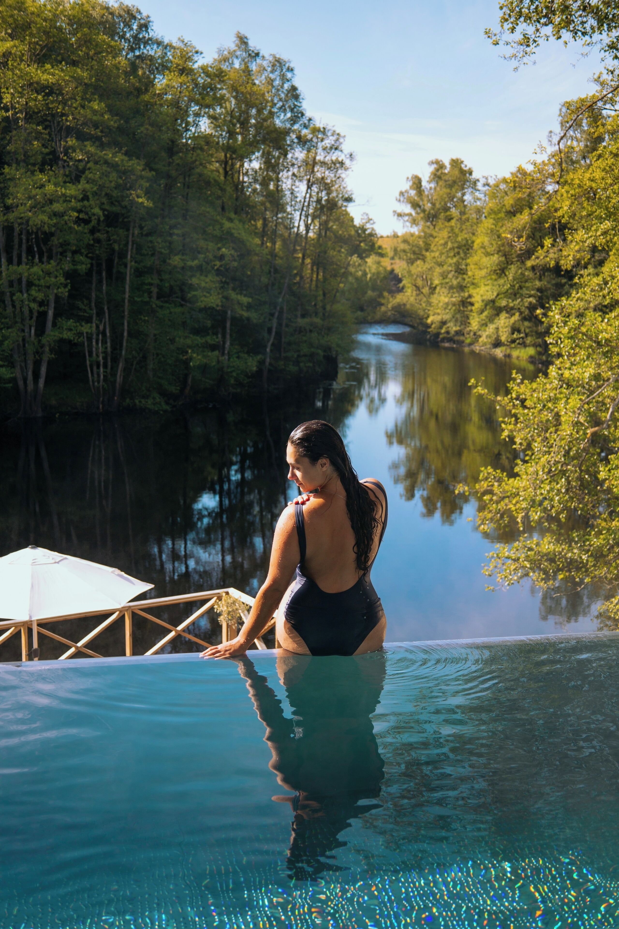 Une femme en maillot de bain est assise au bord d’une piscine à l’eau cristalline, entourée d’une végétation luxuriante.