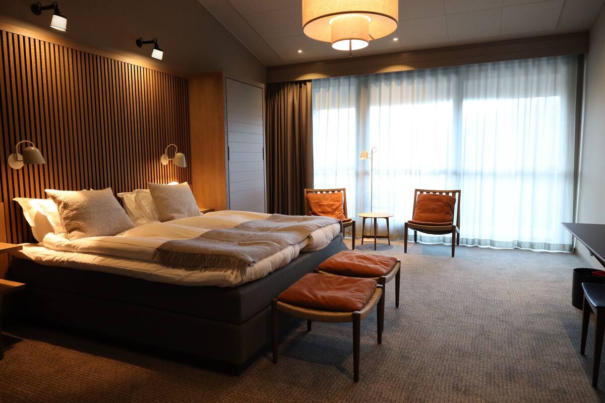 Een hotelkamer met een groot bed, twee stoelen, een klein tafeltje en een groot raam met gordijnen.