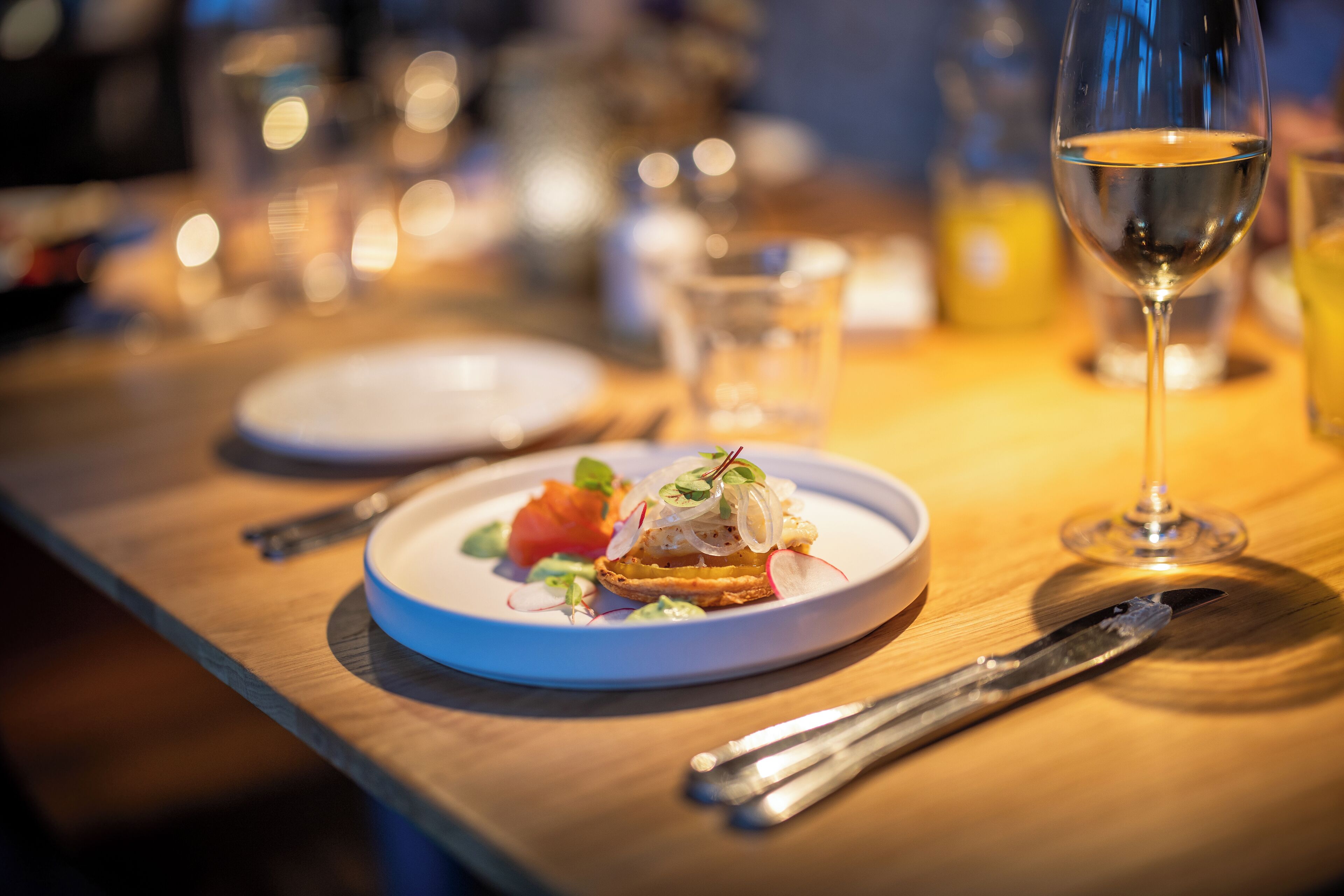A dining table with a plate of food, a glass of white wine, and cutlery.