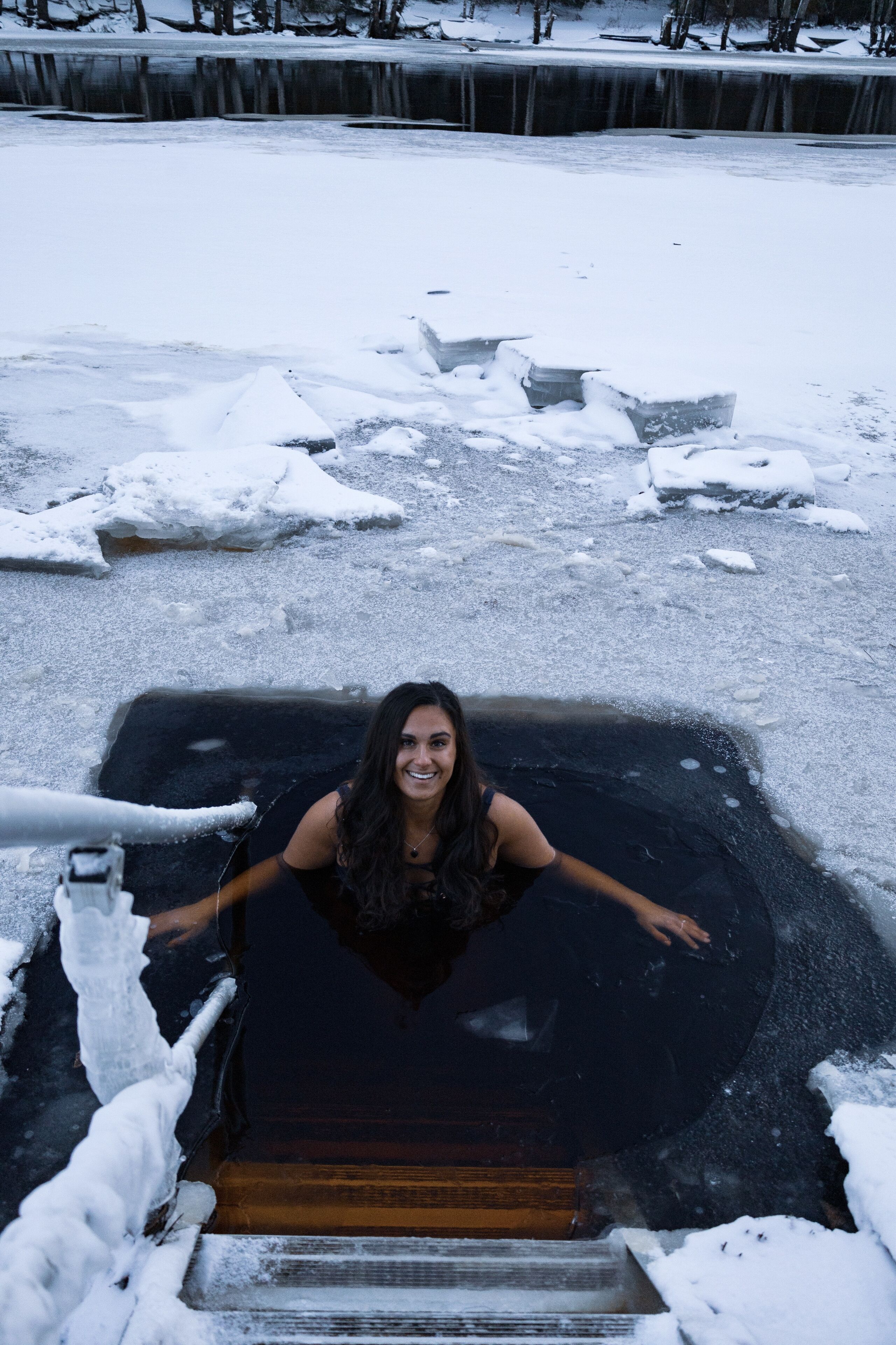 A person in a black swimsuit standing in a partially frozen pool with snow around.