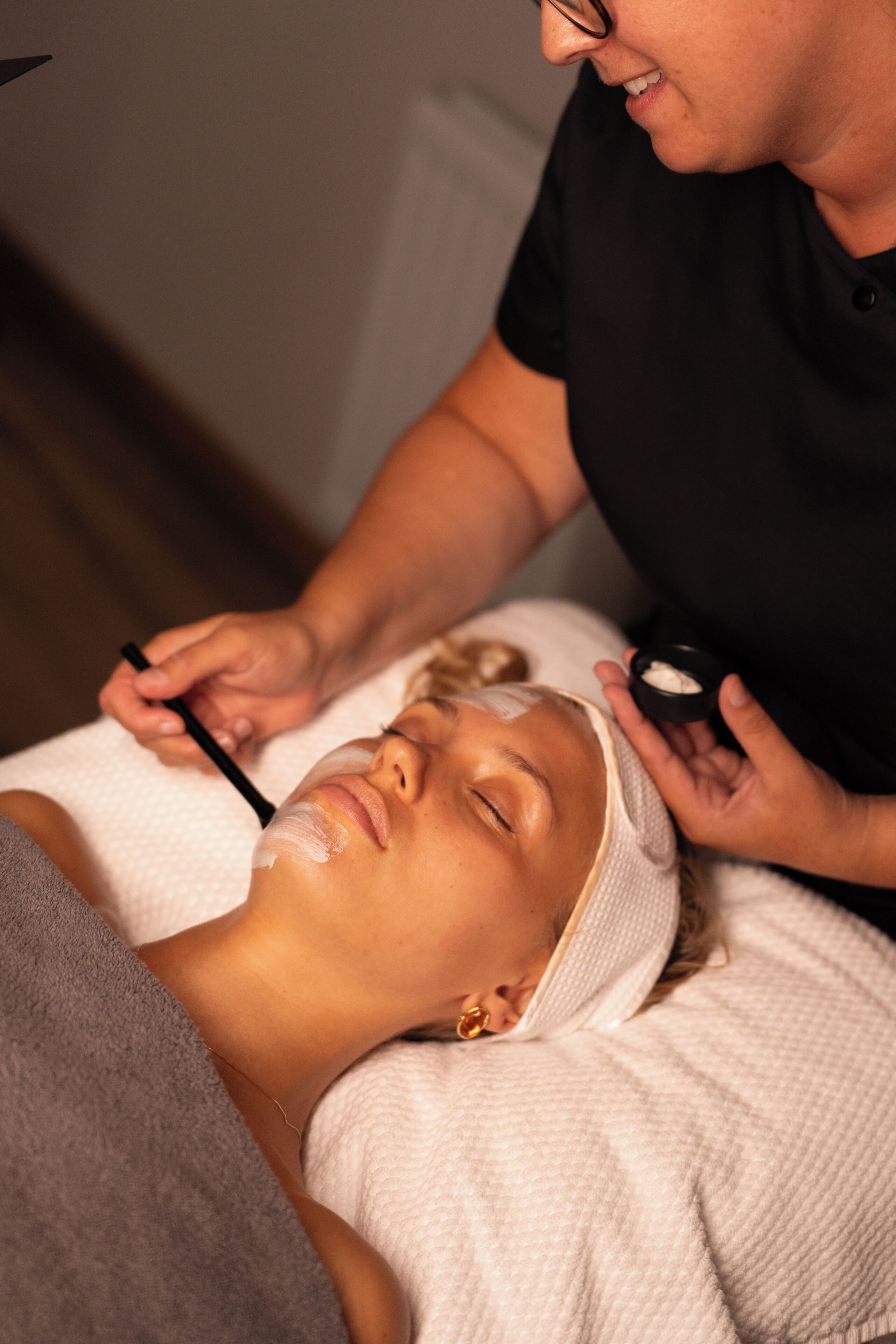 Une personne allongée, une serviette enroulée autour de sa tête, une autre personne appliquant une substance sur son visage, et un petit récipient posé sur la table.