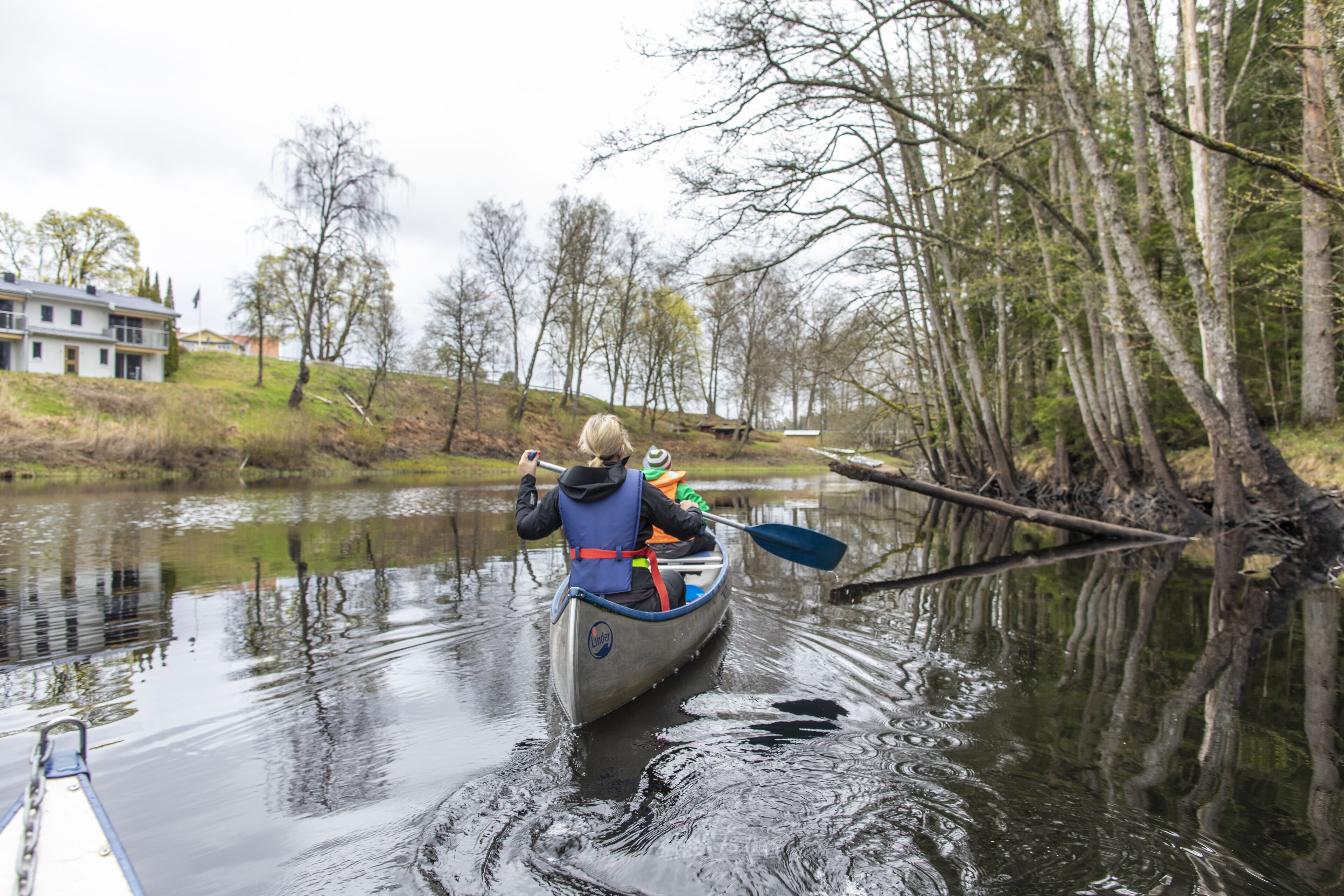 A person in a canoe on a calm river surrounded by trees and a house in the background.