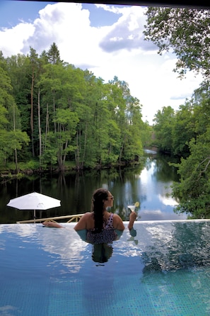 A woman relaxing in a pool with a view of a forested river. - HESTRAVIKEN HOTEL & SPA (Hestra)