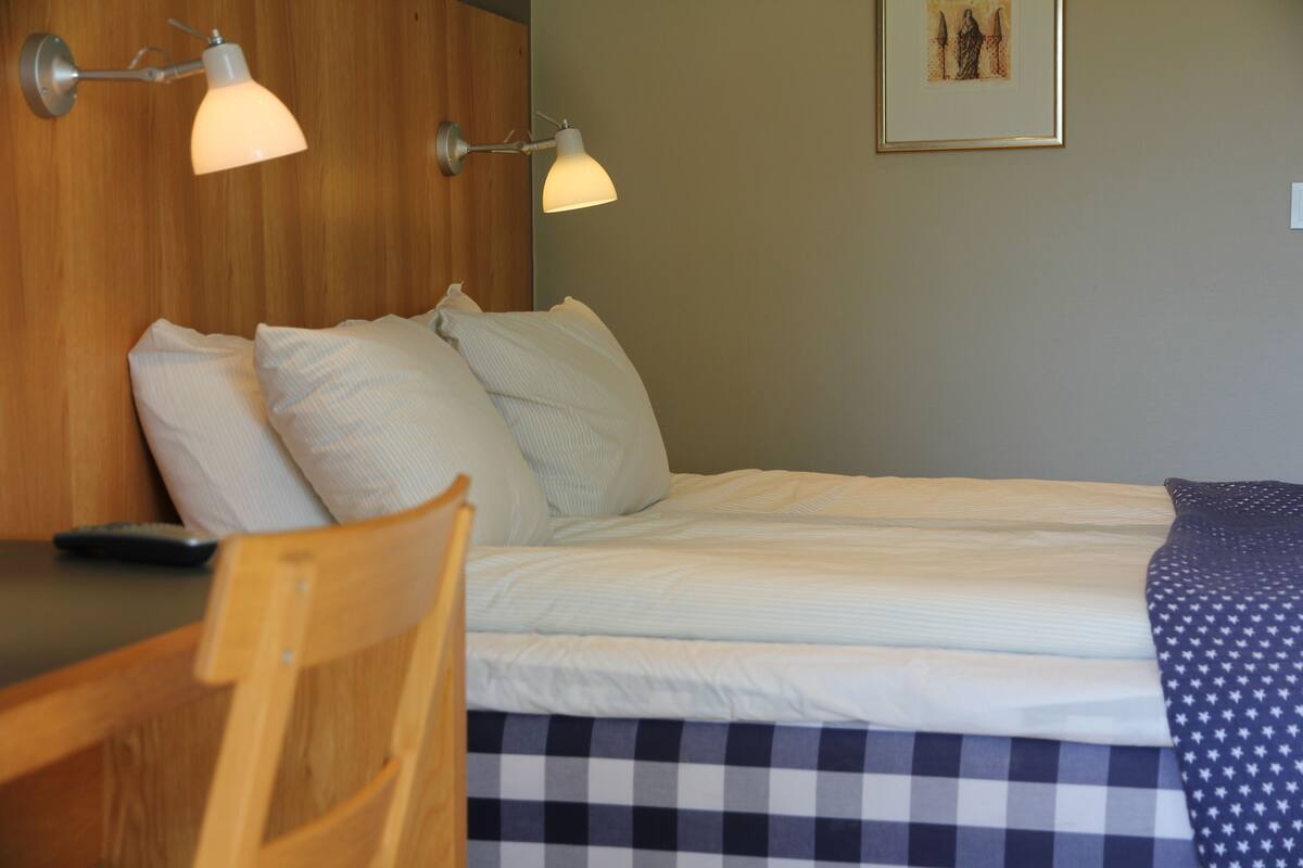 Een net opgemaakt bed met een geruit sprei, twee kussens en een afstandsbediening op een nachtkastje van hout.
