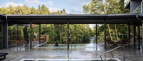 Una piscina exterior con cerramiento de vidrio, rodeada de árboles y una zona cubierta con asientos.