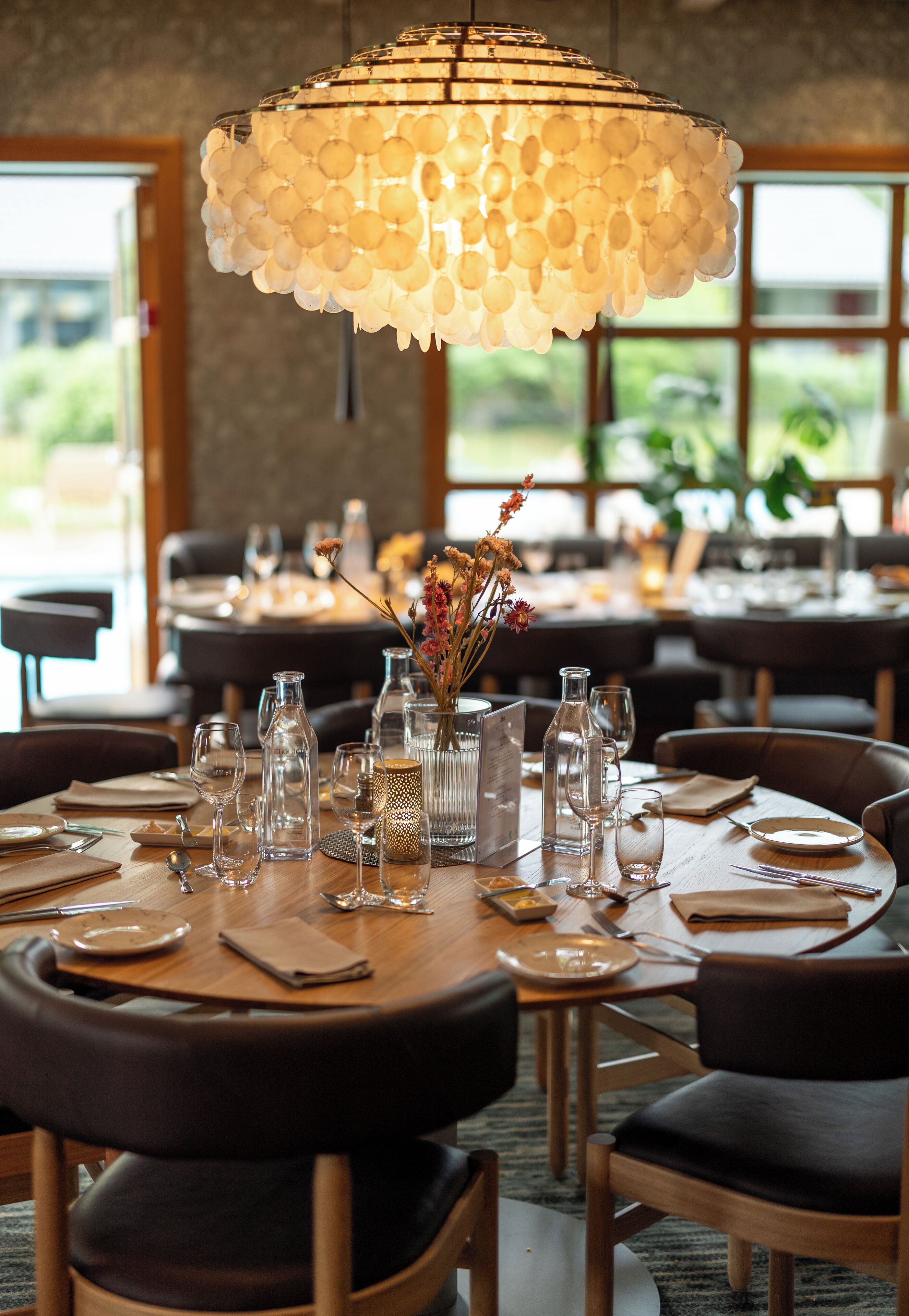 Een restaurantinterieur met ronde tafels gedekt voor dineren, helder glaswerk en een decoratieve hanglamp.