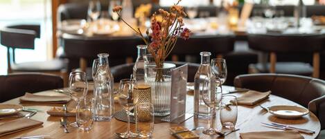 Een restaurantinterieur met ronde tafels gedekt voor dineren, helder glaswerk en een decoratieve hanglamp.