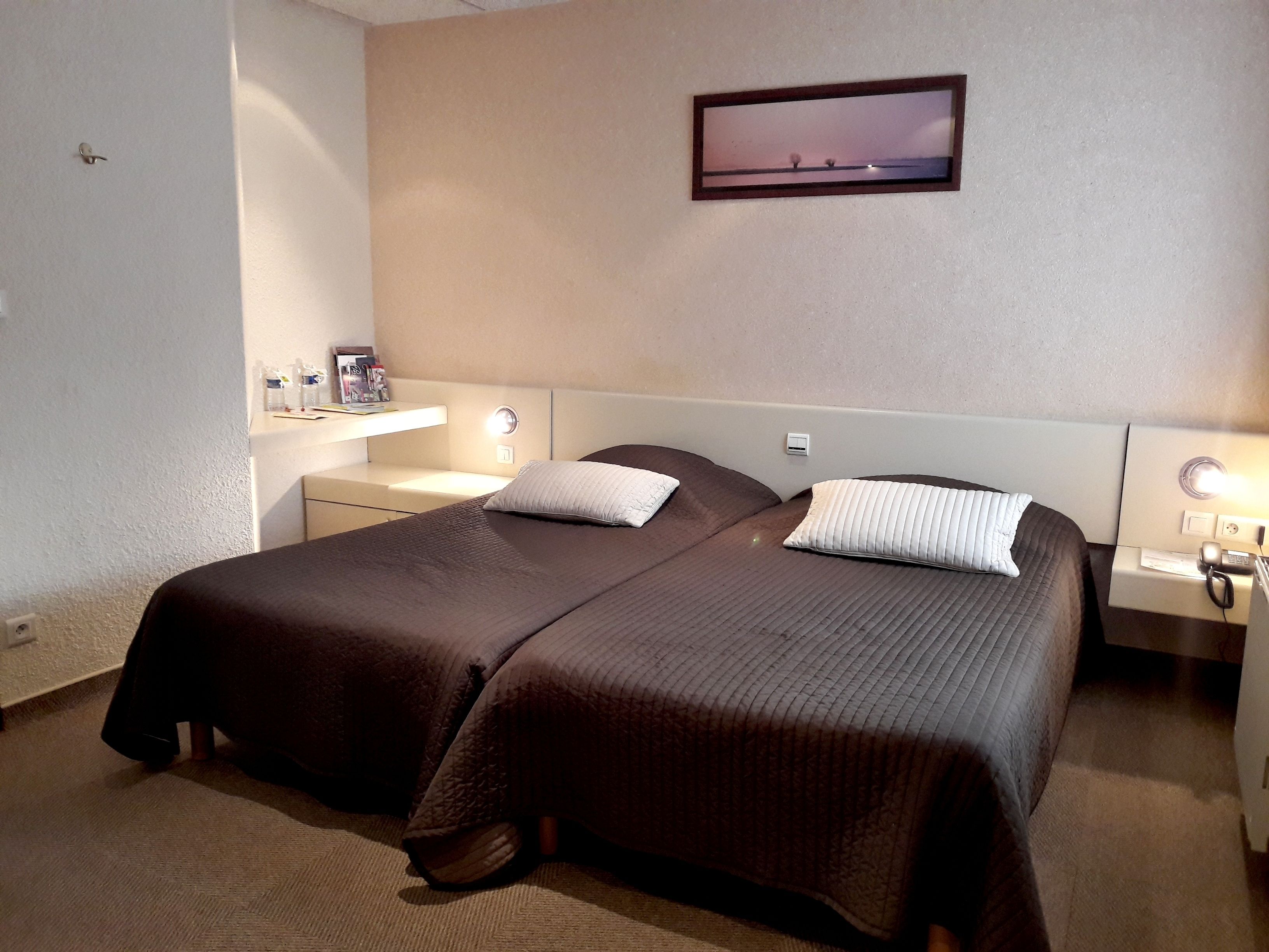 Ein Hotelzimmer mit zwei Betten, einem Nachttisch mit Lampe, einer kleinen Ablage mit Toilettenartikeln und einem gerahmten Bild an der Wand.