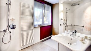 Un bagno moderno con lavandino bianco, doccia con soffione a mano, porta asciugamani e una finestra con tende leggere.