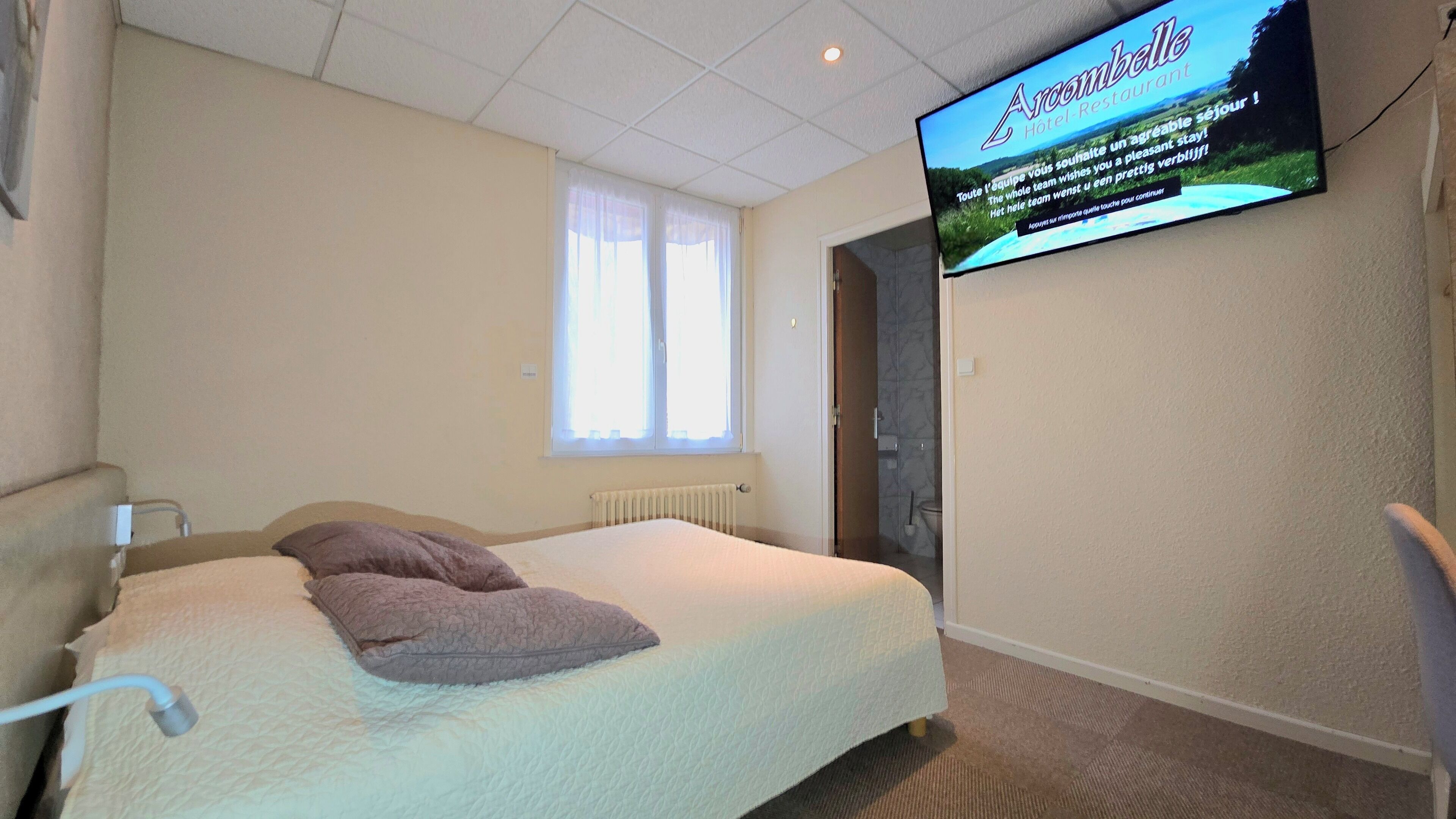 A hotel room with a bed, a TV mounted on the wall, a window with curtains, and a radiator.