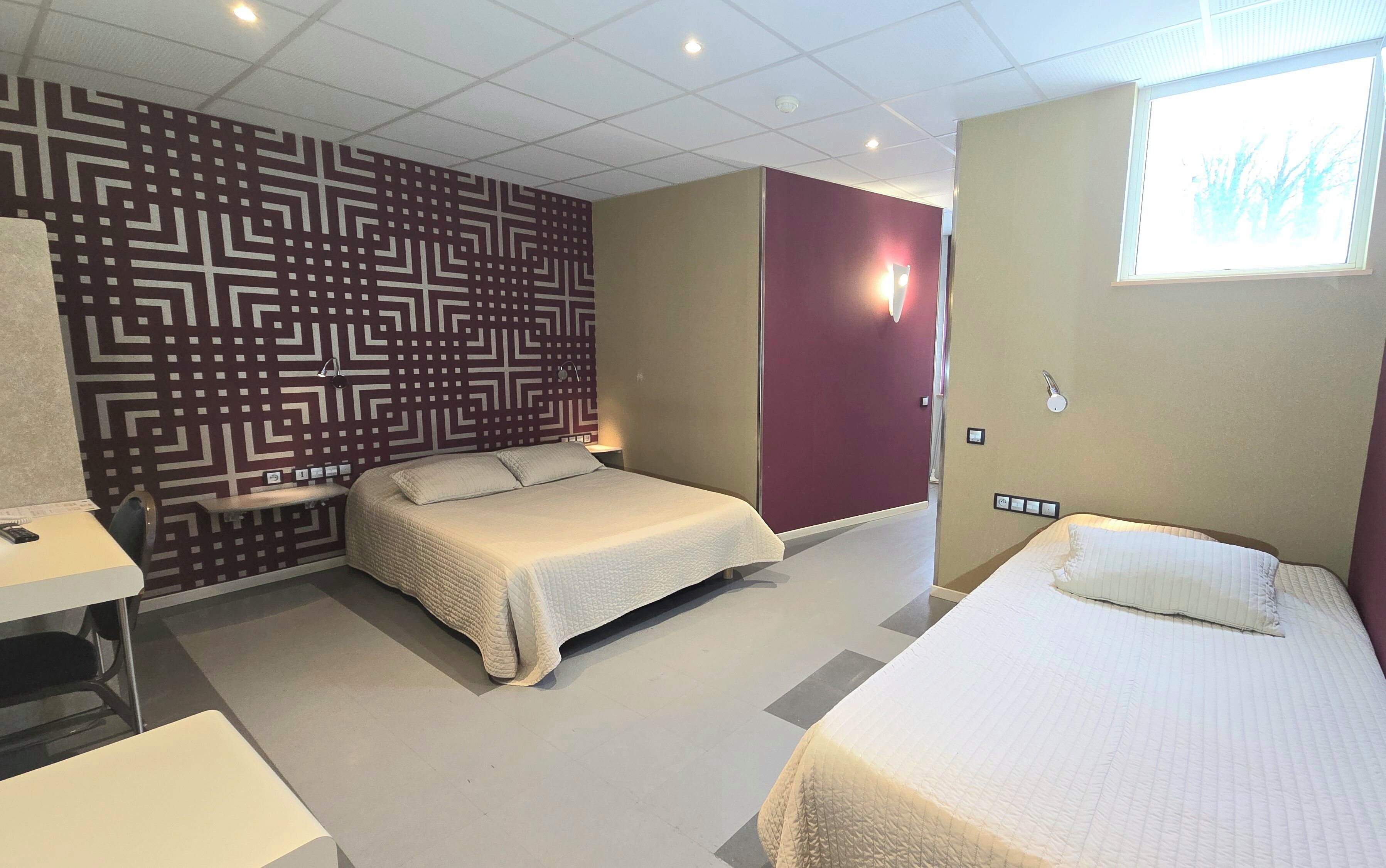 Une chambre d’hôtel avec deux lits, un mur orné de motifs, un mur d’accent rouge et une fenêtre donnant sur un paysage agréable.
