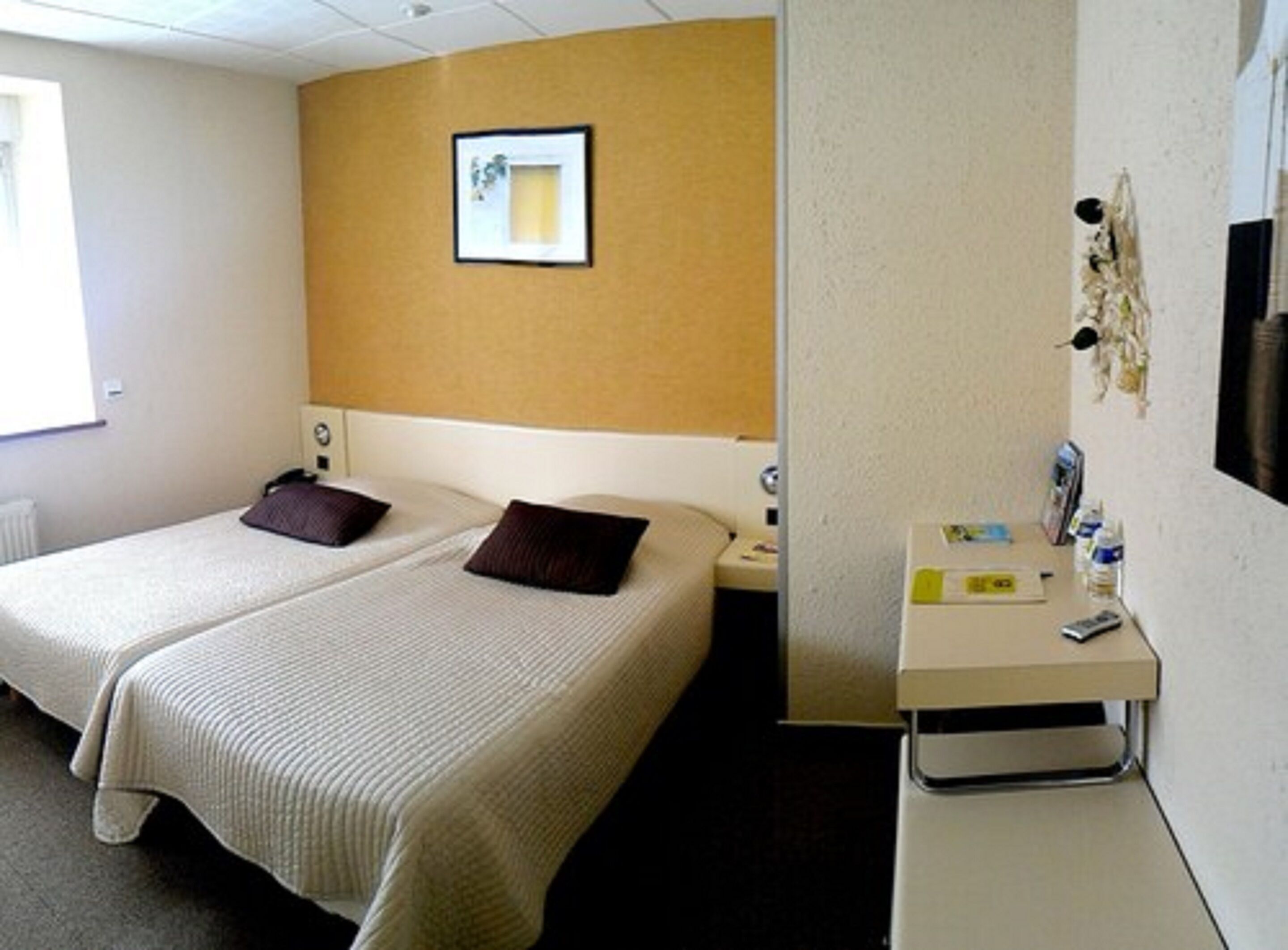 Quarto de hotel com duas camas, uma escrivaninha pequena com cadeira, uma mesinha de cabeceira e uma TV fixada na parede.