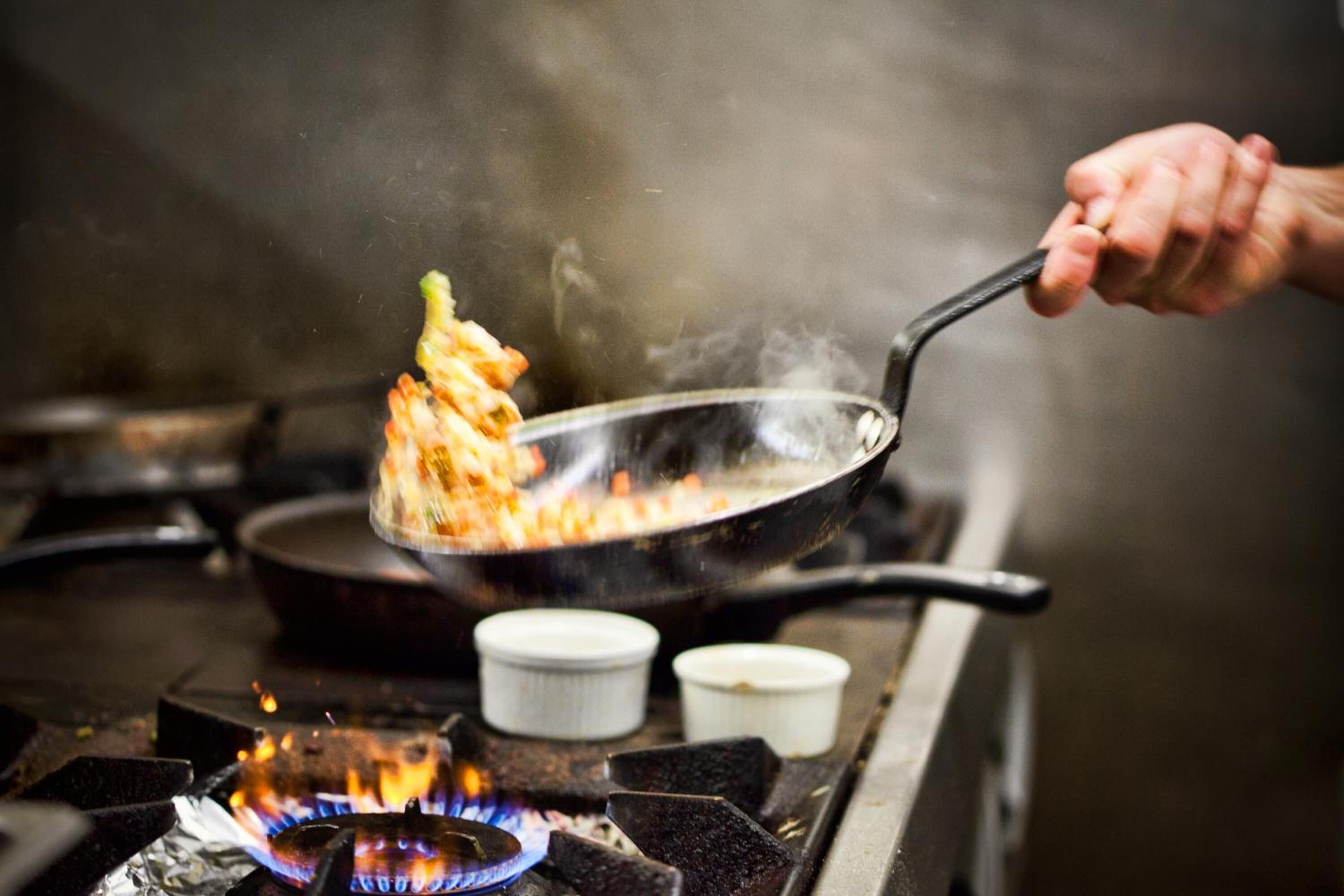 Une main tenant une poêle au-dessus d’une cuisinière où brûlent des flammes.