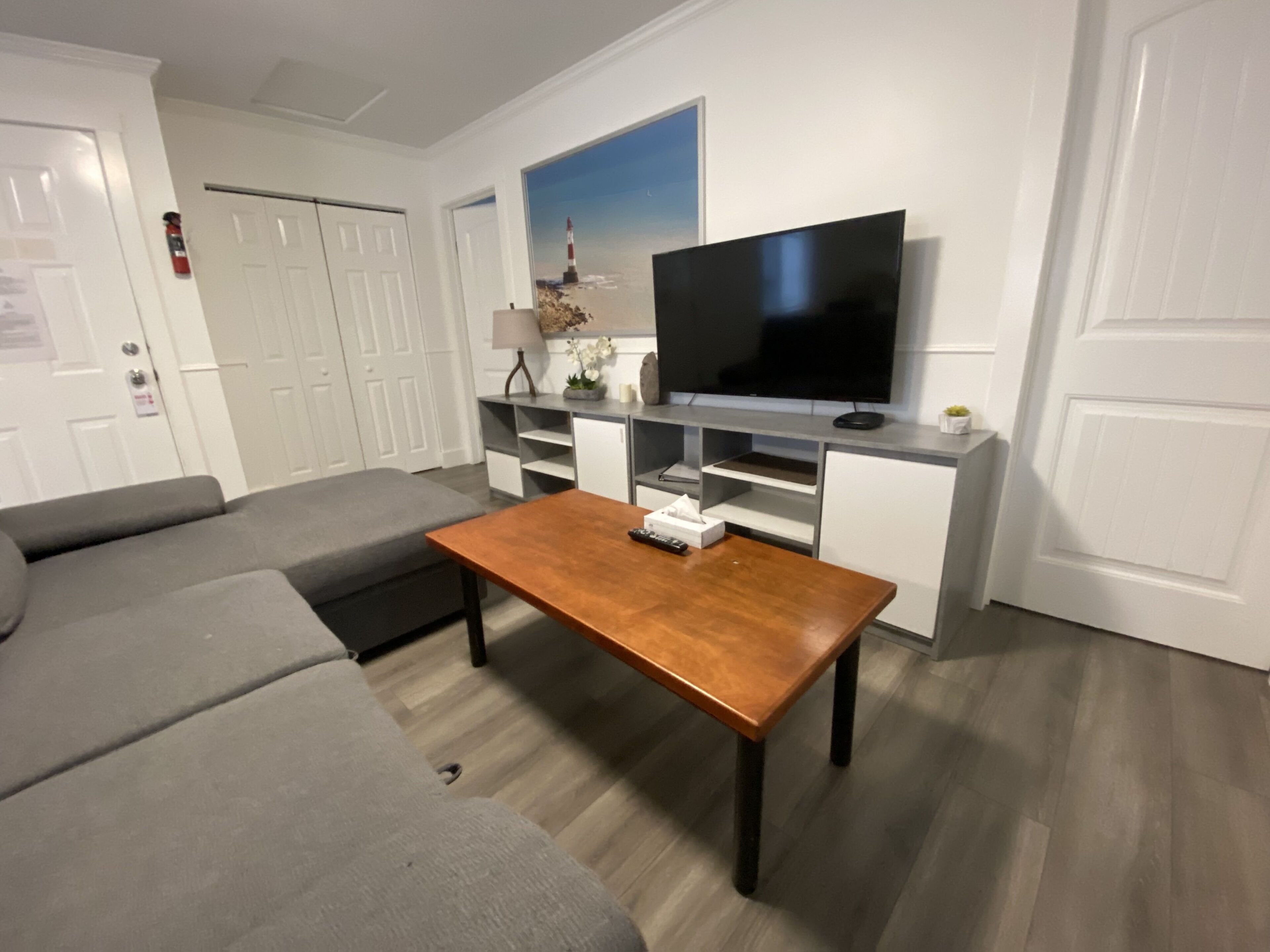 Un salon moderne avec un canapé gris, une table basse en bois, un téléviseur à écran plat posé sur un meuble blanc et un tableau représentant un phare.