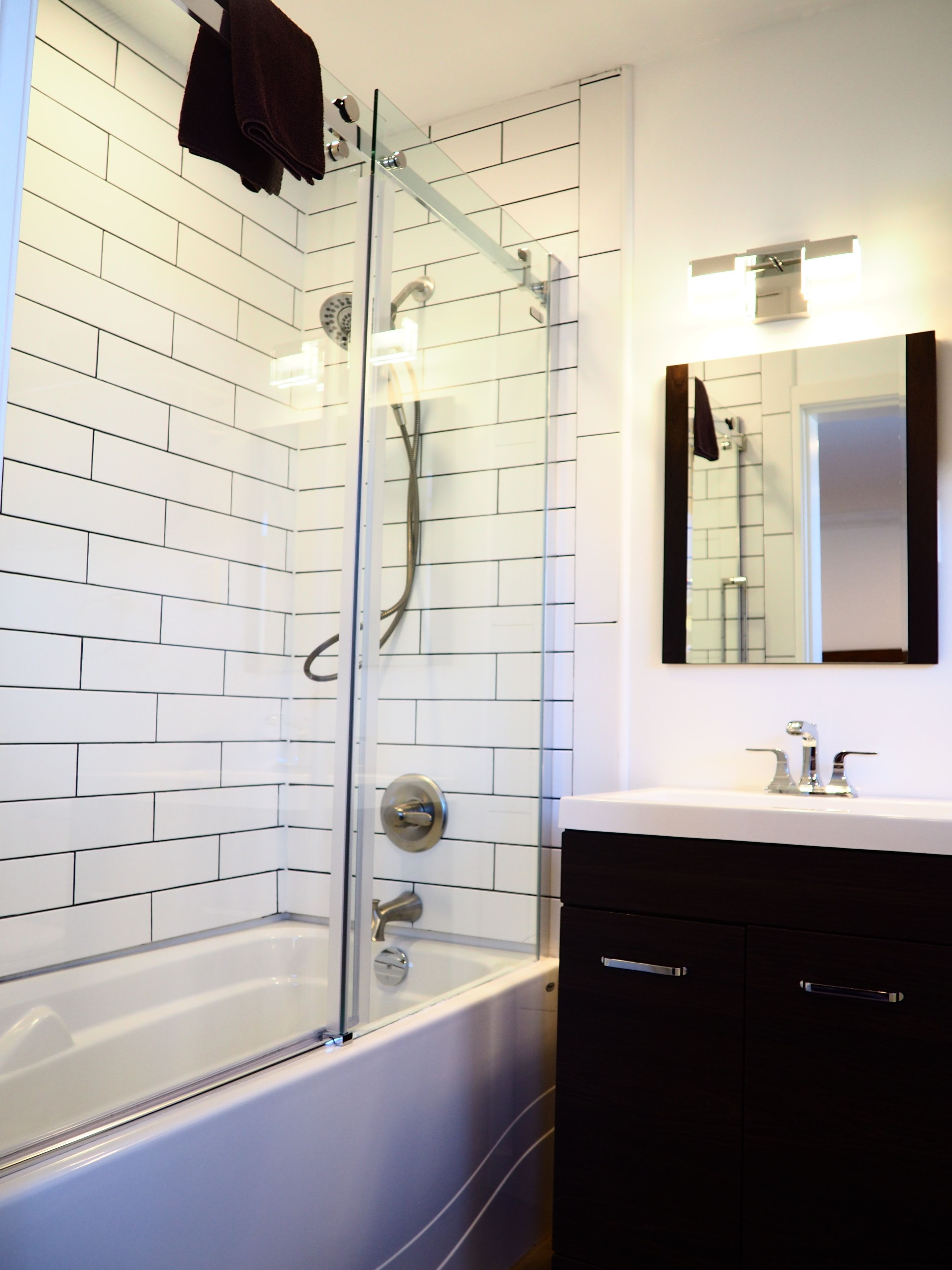 A modern bathroom with a glass-enclosed shower, white tiled walls, a white bathtub, and a black vanity with a mirror.