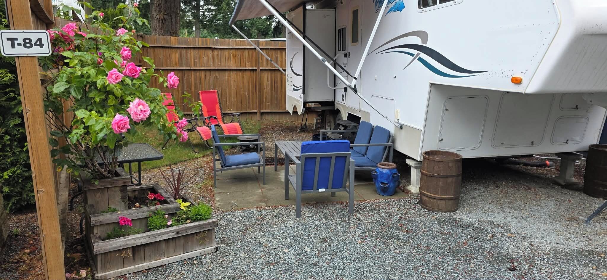 Un véhicule récréatif est stationné dans une cour arrière, qui comprend une terrasse en gravier, une clôture en bois et un panneau indiquant l’adresse T-84.
