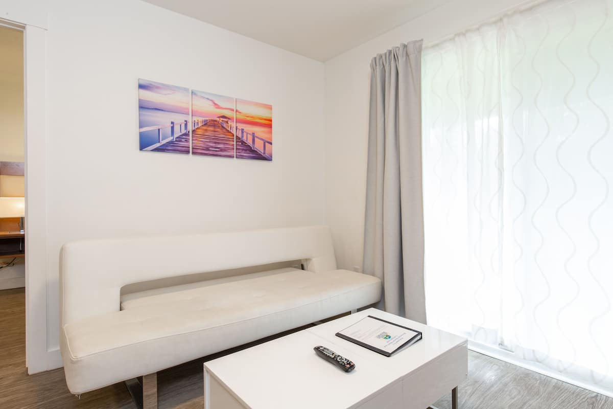 Ein modernes Hotelzimmer mit einem weißen Sofa, einem kleinen Tisch mit einer Fernbedienung und einem Gemälde eines Sonnenuntergangs über einem Steg.