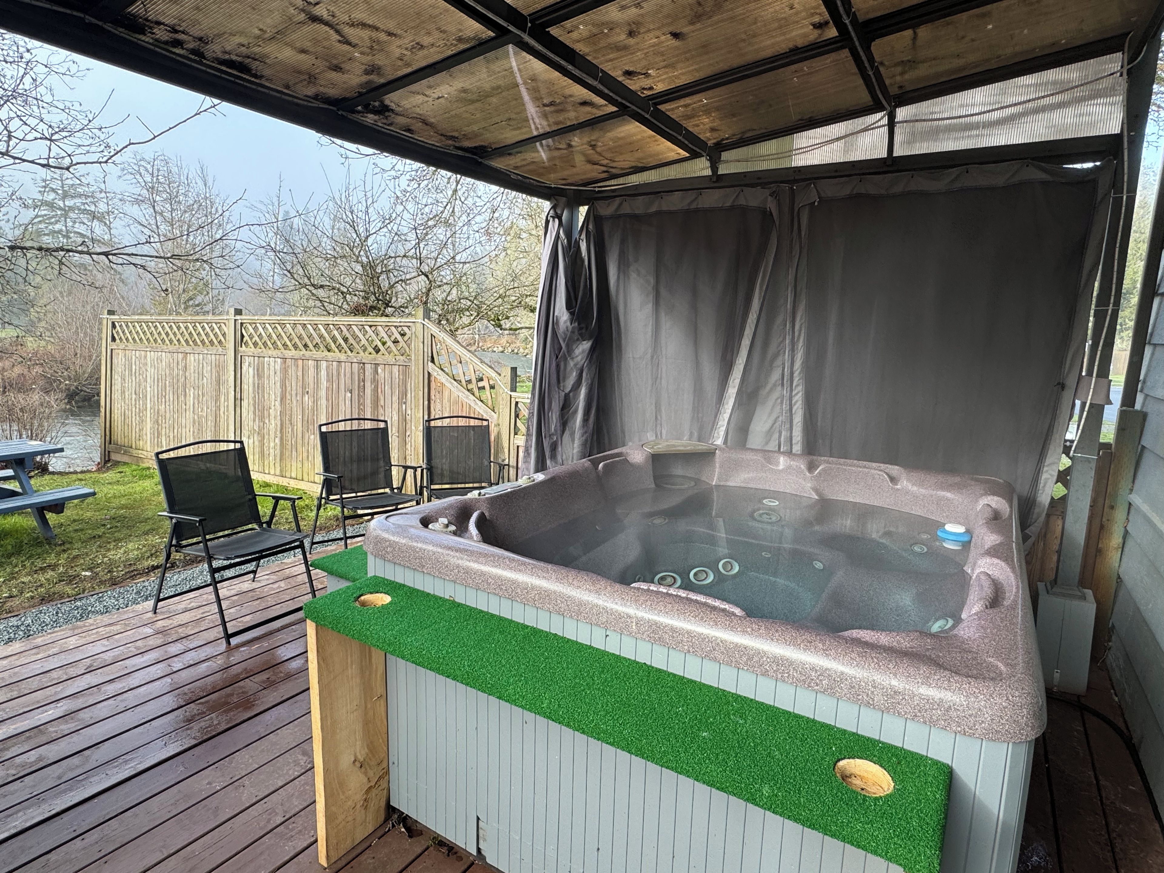 A hot tub with a wooden deck and chairs, a covered area, and a wooden fence in the background.