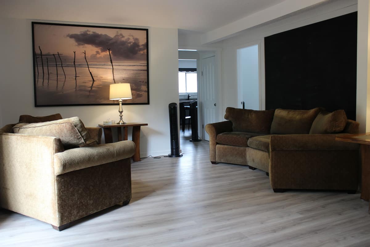 Ein Wohnzimmer mit zwei Sofas, einem Tisch und einer Lampe. An der Wand hängt ein Gemälde und eine Tür führt in einen anderen Raum.