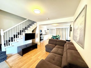 Una sala de estar con una escalera, un televisor de pantalla plana, un sofá y una cocina al fondo.