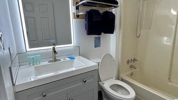 Un baño con lavamanos, espejo, inodoro y ducha. Hay toallas y artículos de aseo en la pared.
