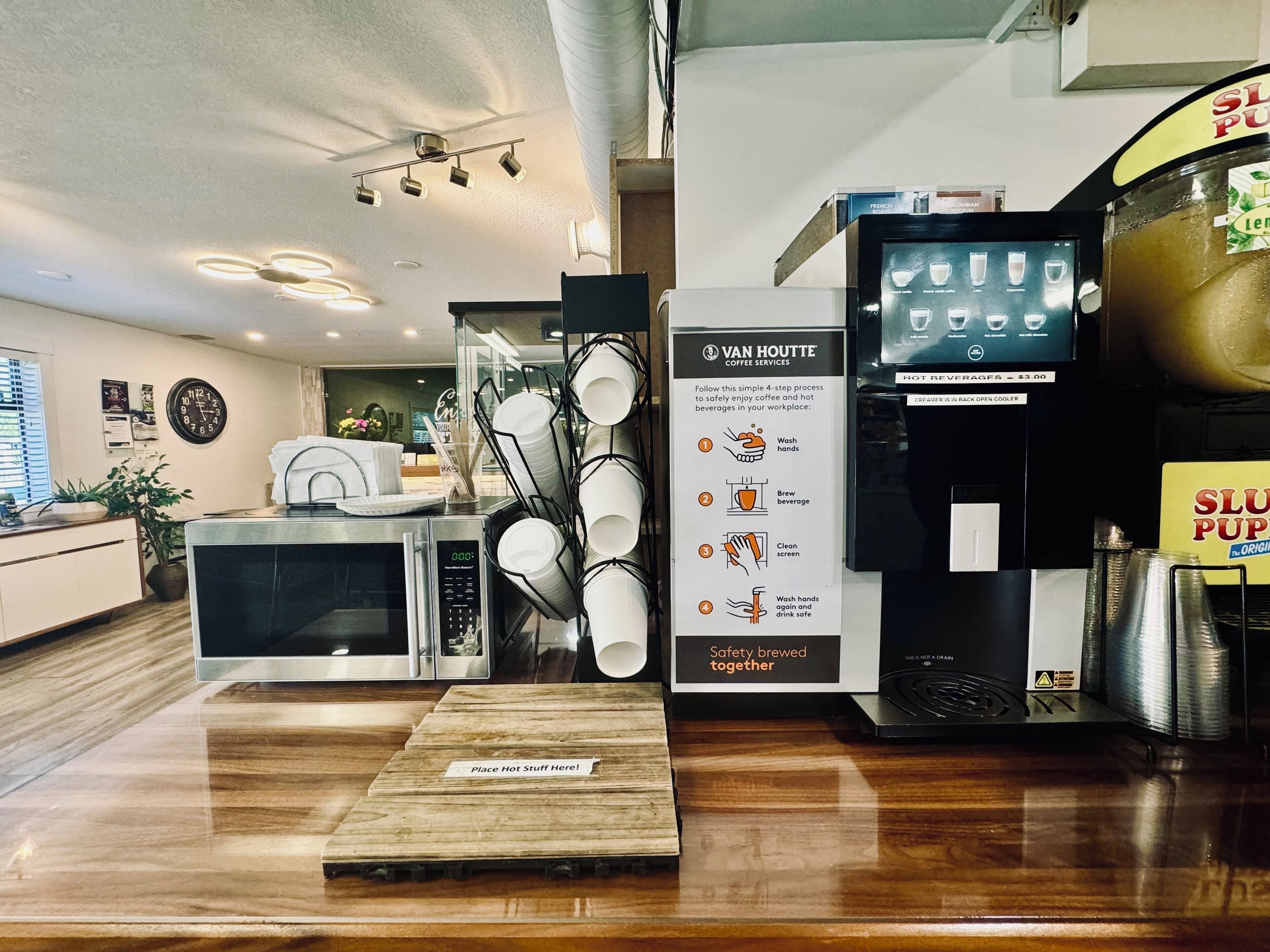 Interior de una cafetería con una máquina de café, un dispensador de bebidas calientes y un letrero que describe los pasos del servicio de café.
