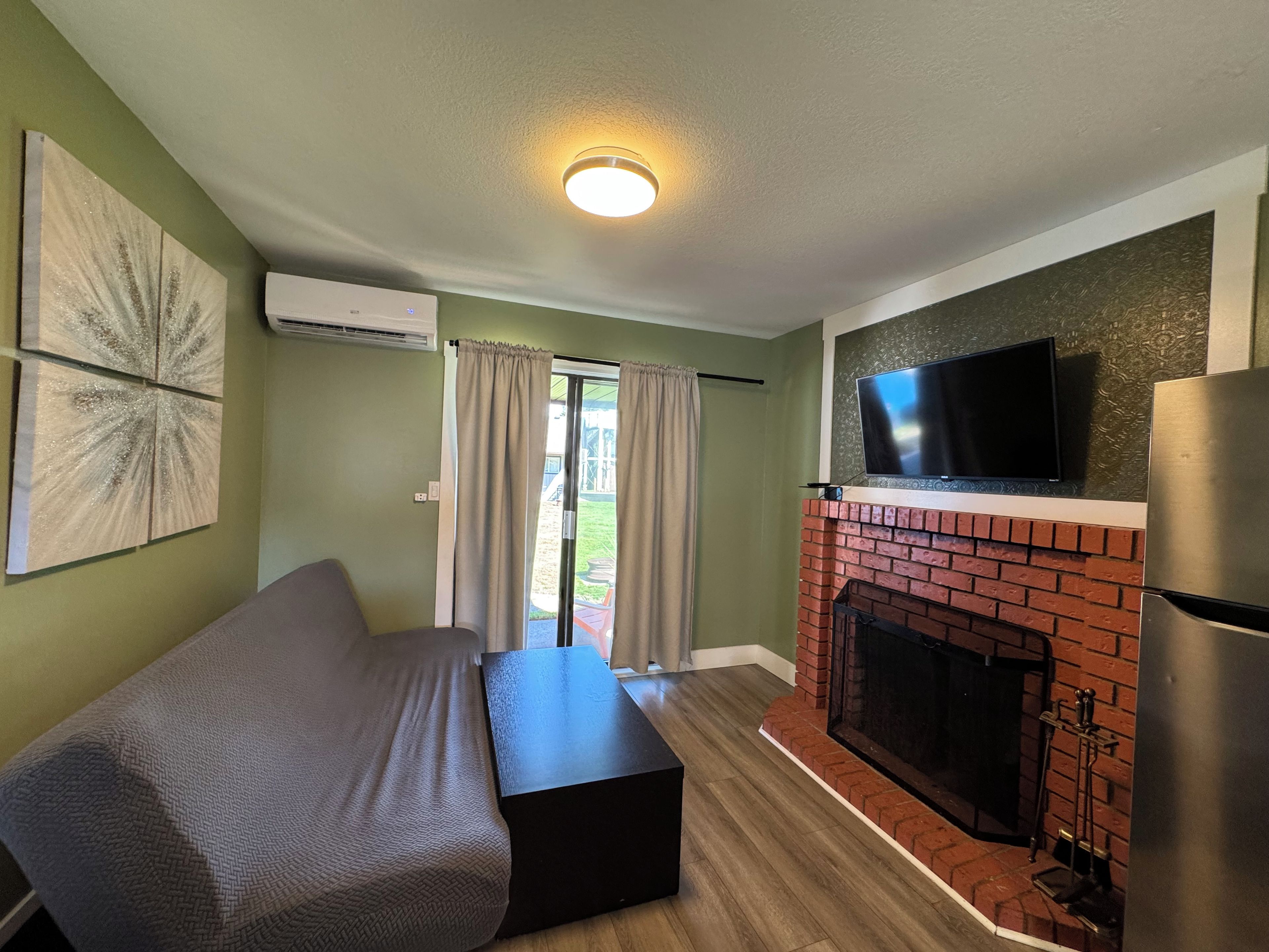 A room with a sofa, a flat-screen TV mounted above a brick fireplace, and an air conditioning unit on the wall.